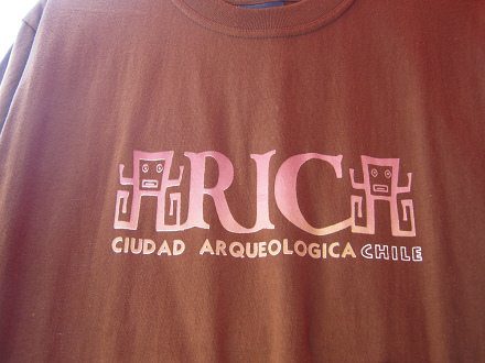 Braunes T-Shirt mit einem
                                Schriftzug "Arica" mit dem A
                                in Form einer menschenhnlichen Figur
                                mit quadratischem Kopf mit rechteckigen
                                Lippen und Knopfaugen, und ohne Rumpf